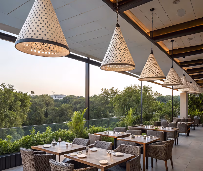 Outdoor restaurant seating area with wooden tables, wicker chairs, and hanging cone-shaped lights overlooking green trees.